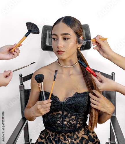 Young woman with sleek hair and elegant makeup sits on chair against white background while several hands apply powder, blush, mascara, and lip color with cosmetic brushes in studio portrait