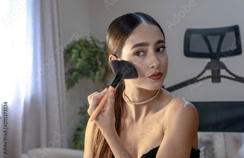 Young woman applies powder makeup with large brush during personal beauty routine, seated indoors beside bright window light with blurred plant and chair background, calm elegant expression