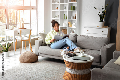 Young African-American woman reading magazine on sofa at home
