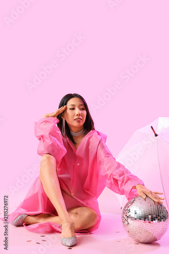 Fashionable young Asian woman in raincoat with disco ball and umbrella sittin...