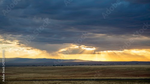 Dramatic golden sunbeams bursting through dark storm clouds over vast open prairie landscape at sunset with rain shafts in Wyoming