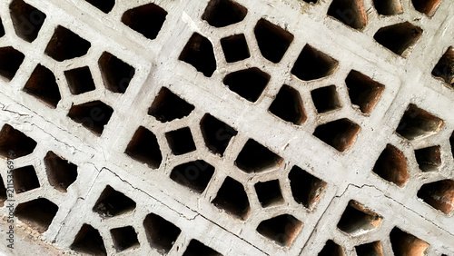 Concrete ventilation blocks arranged diagonally, forming a geometric pattern with hollow openings. Rustic architectural texture featuring aged cement and exposed red clay bricks, ideal as an abstract 