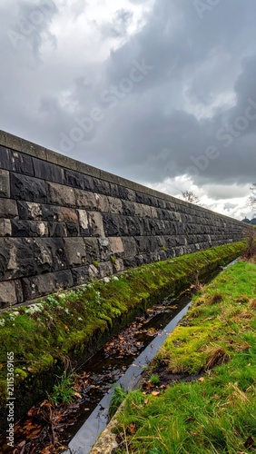 Long stone wall beside a narrow, overgrown channel under cloudy sky