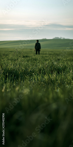 Person standing in a lush green field during early morning light, surrounded by tall grass and a serene landscape.