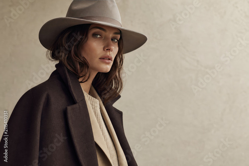 Elegant woman in neutral-toned wide-brim fedora and long wool coat posing confidently against minimalist beige backdrop, timeless fashion portrait with soft natural lighting