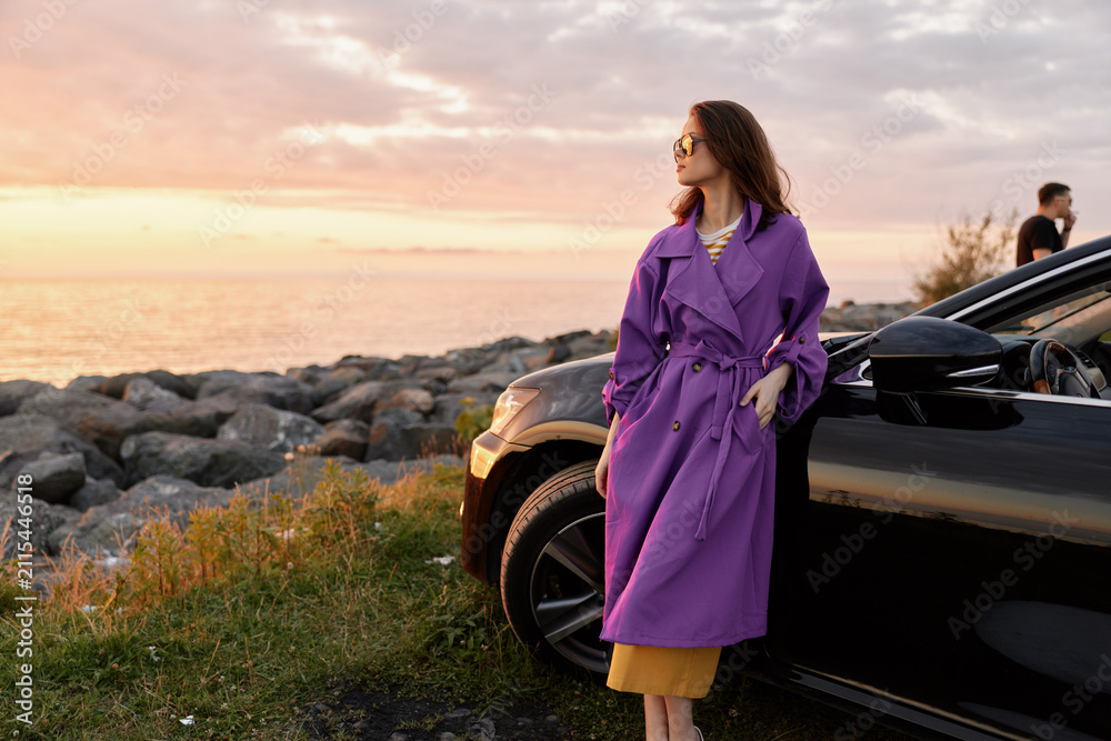 Fototapeta premium A woman in a purple coat stands beside a dark car at sunset, gazing toward the horizon with a confident pose as warm light reflects off rocks and calm water nearby.