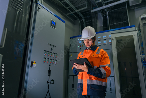 Professional Asian industrial engineer in safety uniform using a digital tablet with stylus to record maintenance data in front of an electrical capacitor bank control panel in a factory.