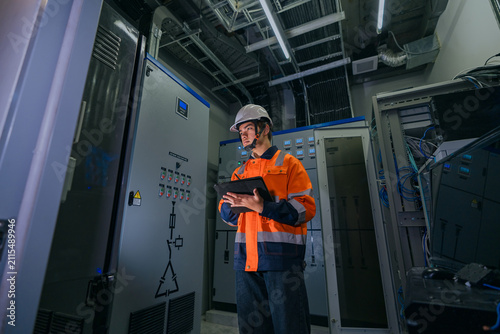 Professional IT network engineer or electrical technician inspecting server rack and industrial power control system using a digital tablet in a modern manufacturing data facility.