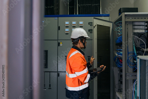 Professional IT network engineer in safety uniform holding digital tablet inspecting ethernet cables and server rack in a modern industrial data center communication room.