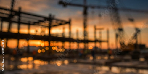 A blurred construction site at sunset, featuring cranes and scaffolding. The warm tones of the sunset reflect off wet surfaces, creating a dynamic atmosphere.