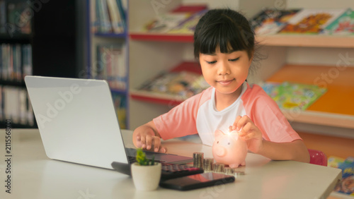 Asian girl putting coin into piggy bank while using laptop for online banking learning.