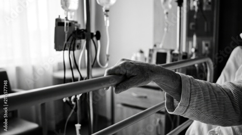Elderly Hand Gripping Hospital Bed Rail With IV Drip In Background