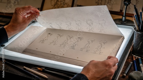 Hands flipping animation sheets on a lightbox showing pencil-drawn character walk cycle with pencils and studio workspace tools