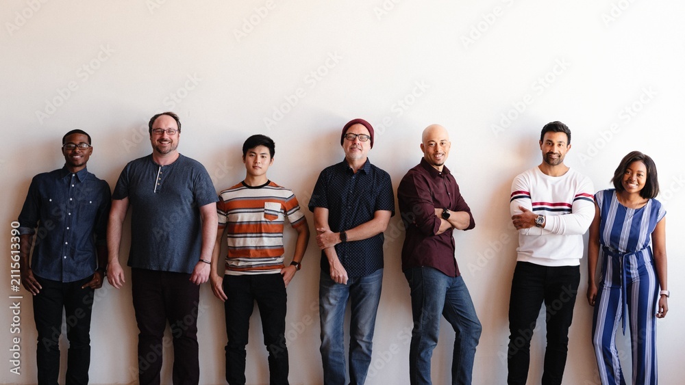 Obraz premium A diverse group of seven people standing against a white wall, smiling.