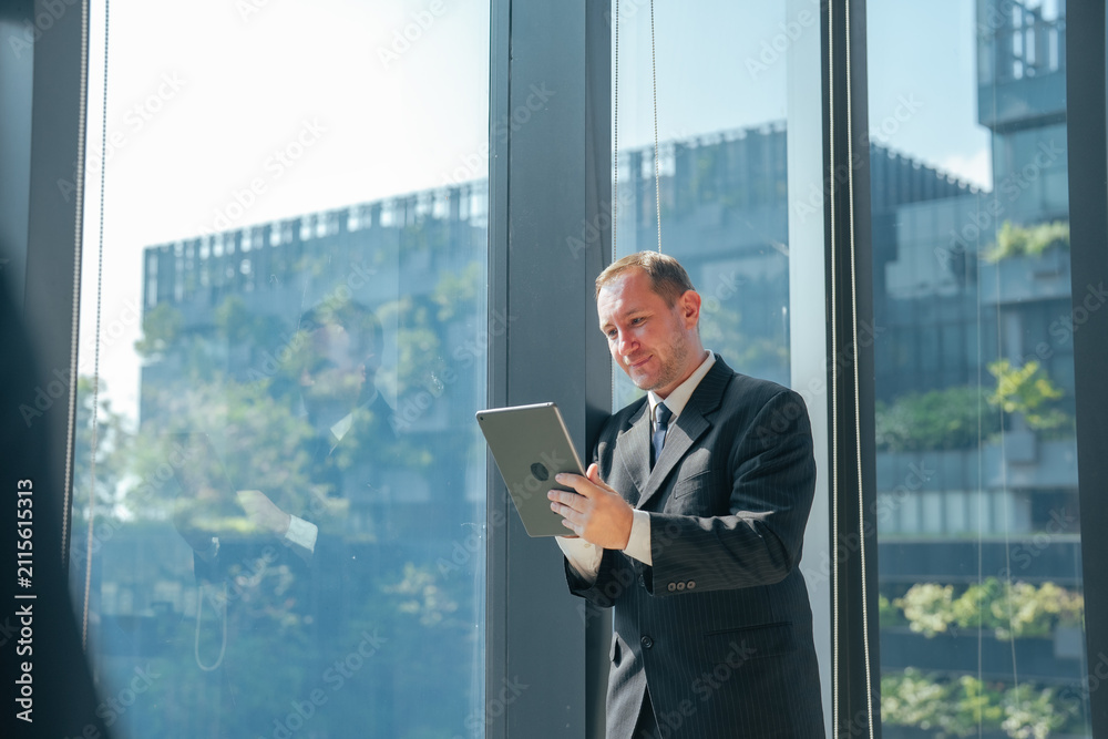 Fototapeta premium Businessman reading tablet in modern office with large window. Professional reviewing business data, planning work, managing projects and using digital technology in workplace.
