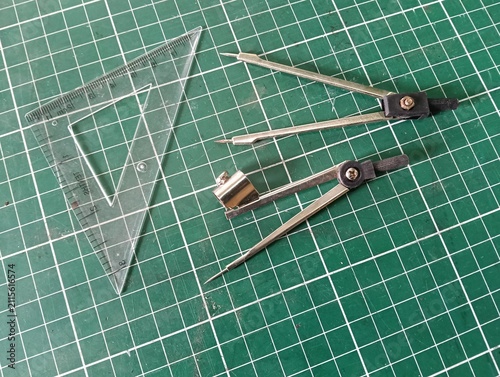 Technical drawing tools including a metal drawing compass, transparent triangular ruler, and measuring instruments arranged on a green cutting mat, used for geometry, drafting, engineering, and precis