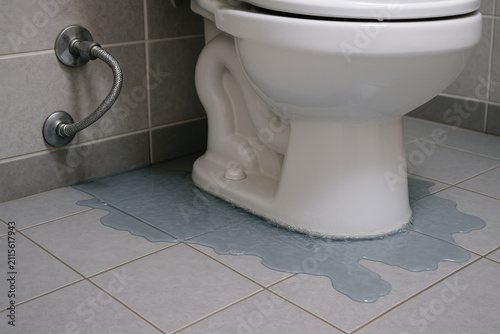 Toilet leaking water onto tiled bathroom floor, plumbing disaster concept close up, water damage visible around base, indoor restroom emergency