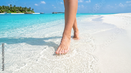 Close perspective showing female bare feet splashing lightly through warm transparent tropical sea waves washing across pristine pure white sand island shoreline