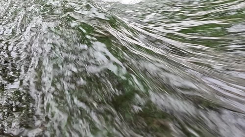 Fast-moving stream water rushes steadily over green grass and algae underwater, creating streaked white ripples that flow diagonally across the frame in a natural rocky creek.