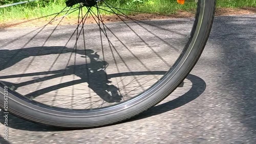 The bike's front wheel spins quickly, silver spokes and orange reflector sparkling in the sunlight as you ride along a forest road bordered by green grass.
