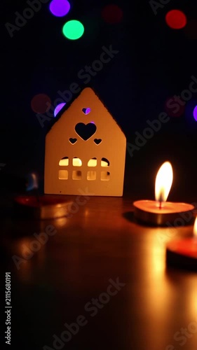 Heart cutout house lantern sits on a table as tealight candles are lit one by one across frames. Each new flame flares, steadies, and warms the scene while colorful bokeh lights shimmer behind.