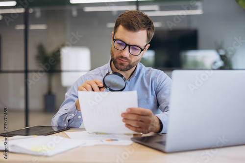 Man using magnifying glass to inspect documents in office, focusing on details and analysis during business review