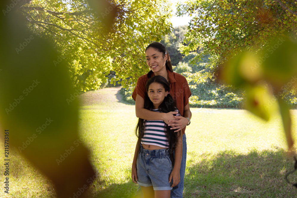 Obraz premium African American mother embracing youth daughter in park under large tree, wearing rust shirt