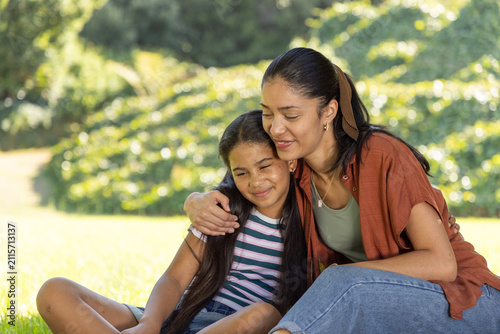 African American mother and youth daughter sitting close on sunny grass in park, showing necklace