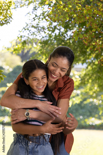 African American mother and youth daughter hugging under tree in park showing rust top and watch