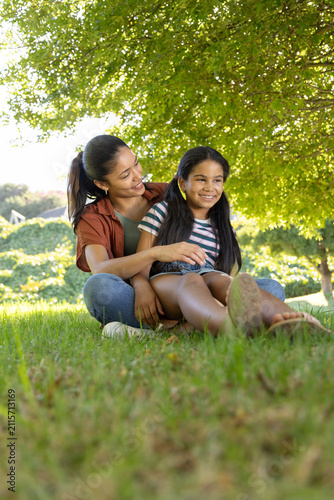 African American mother and child sitting on grass under tree, hugging, bare feet, white sneaker