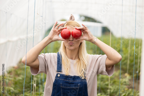 Young woman having fun, holding ripe tomatoes over eyes, making a funny face in a farm greenhouse