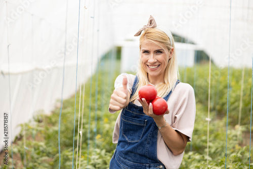 Happy woman farmer showing two fresh organic tomatoes and thumb up in a sunny commercial greenhouse