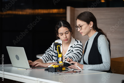 Robotics Education with a female teacher and Asian student using a laptop beside a yellow robotic arm and electronic equipment in a STEM classroom