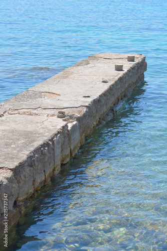 Foto An old weathered concrete pier with small stone bollards stretches diagonally into the calm clear turquoise sea water on a sunny day