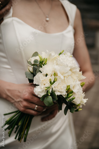 A slender, thin, model-looking bride in a white fitted dress with a precious addition on her neck holds a bouquet of white roses made by florists in her hands