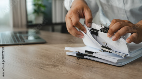 Hands sort clipped paper documents beside laptop on wooden desk, organizing pages and reviewing file bundles for office records management, document handling, filing workflow with copy space.