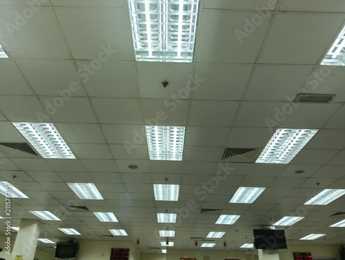  view of a commercial office ceiling featuring multiple recessed fluorescent LED light fixtures, suspended ceiling tiles, air conditioning ventilation grilles, and mounted display monitors. The interi