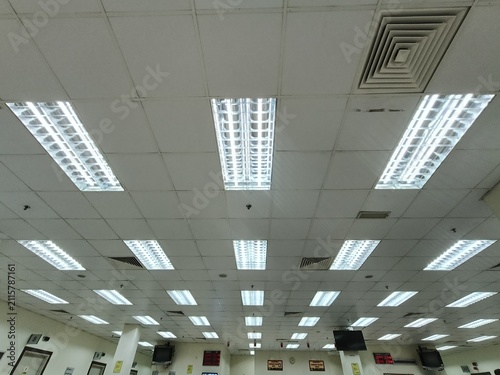  view of a commercial office ceiling featuring multiple recessed fluorescent LED light fixtures, suspended ceiling tiles, air conditioning ventilation grilles, and mounted display monitors. The interi