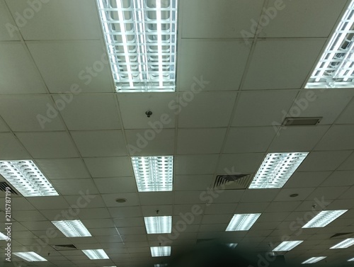  view of a commercial office ceiling featuring multiple recessed fluorescent LED light fixtures, suspended ceiling tiles, air conditioning ventilation grilles, and mounted display monitors. The interi