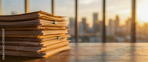 A stack of documents on an office desk with a city skyline in the background at sunset