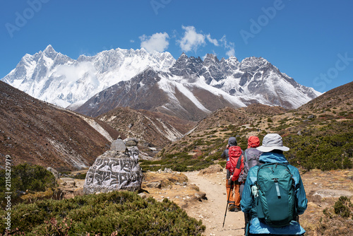 Hikers trekking on Everest Base Camp trail walking past sacred Mani prayer stone against majestic Himalayan mountains background, Nepal