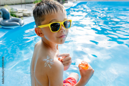 Little boy applying sunscreen in sun shape on his shoulder near pool.