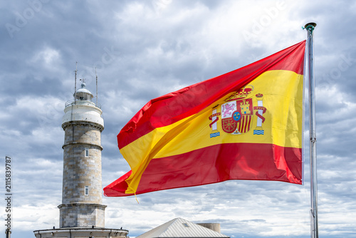 Spanish flag waves proudly near a historic lighthouse under a cloudy sky.