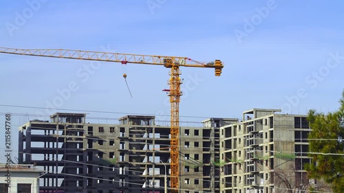 Video footage from a construction site where a yellow tower crane is being used to erect a multi-story building. The raised hook and free slings swing, and the boom swings toward the loading area