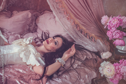 Bild auf Leinwand Sensual dark-haired lady posing on a velvet bed surrounded by blooming pink peon