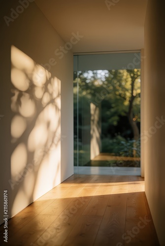 Warm sunlight streams through a modern hallway onto a wooden floor, casting tree shadows on the wall. Use: architectural design, interior decor, li...