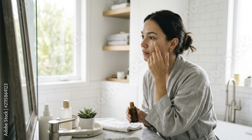 Young woman gently applies topical facial plumping serum to her face, reflecting healthy skin during a serene morning beauty routine in a modern bathroom.