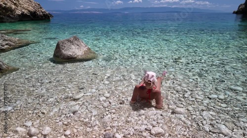 Woman sitting on pebble beach with feet immersed in serene Adriatic Sea. Enjoying a summer vacation in a hidden cove surrounded by natural rocky cliffs under bright blue sky in Cres Island of Croatia