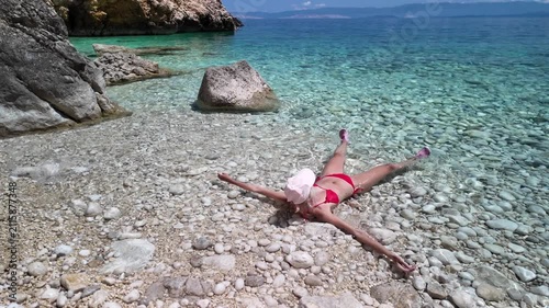 Woman sitting on pebble beach with feet immersed in serene Adriatic Sea. Enjoying a summer vacation in a hidden cove surrounded by natural rocky cliffs under bright blue sky in Cres Island of Croatia