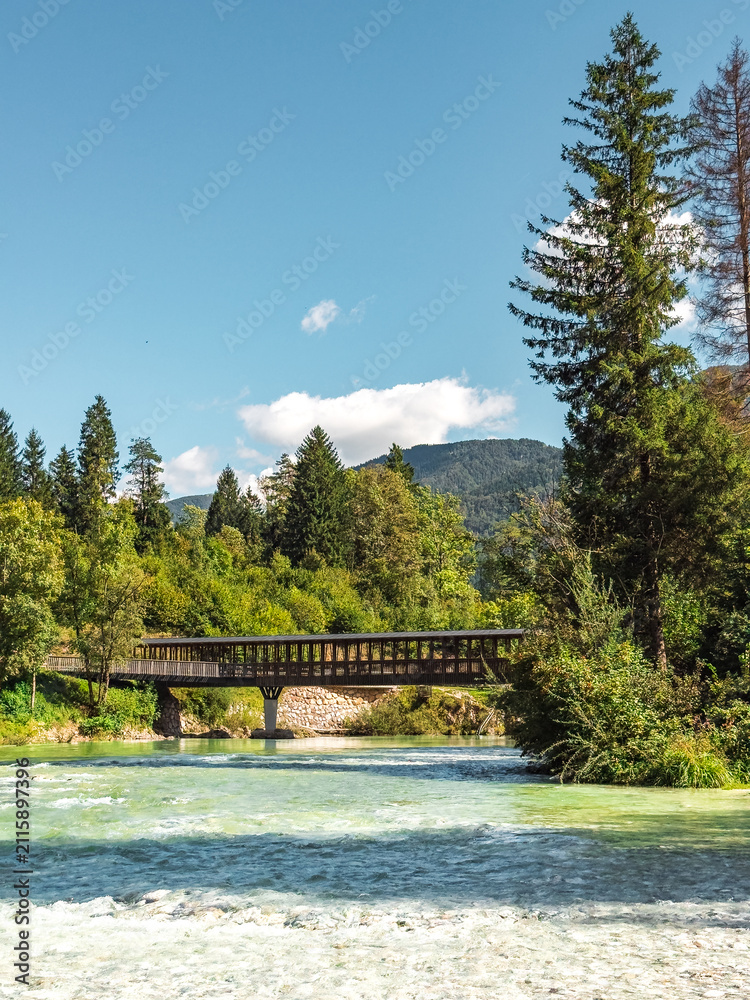 Fototapeta premium Wooden pedestrian bridge crossing the turquoise waters of the Sava Bohinjka River surrounded by lush forest and alpine scenery
